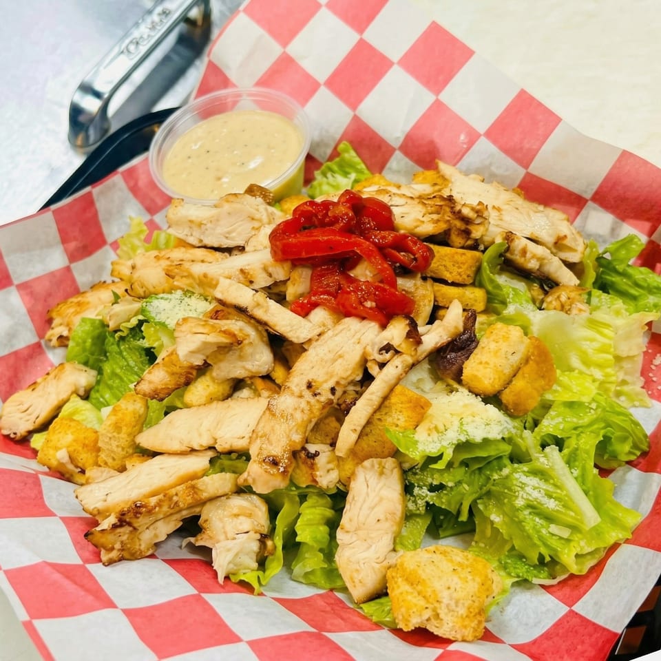 Grilled chicken Caesar salad with red peppers and dressing in a red and white checkered basket.