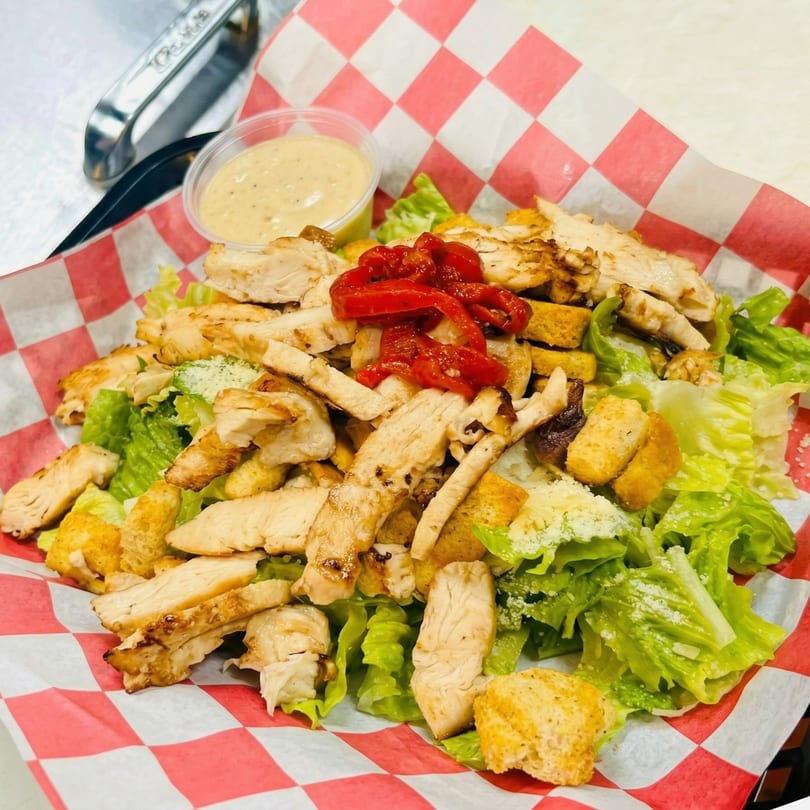 Grilled chicken Caesar salad with red peppers and dressing in a red and white checkered basket.