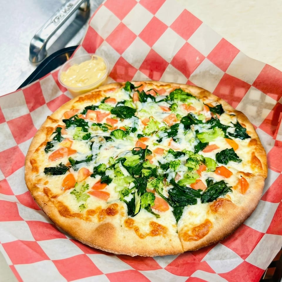 A white pizza with broccoli, spinach, and diced tomatoes, served on red and white checkered paper.