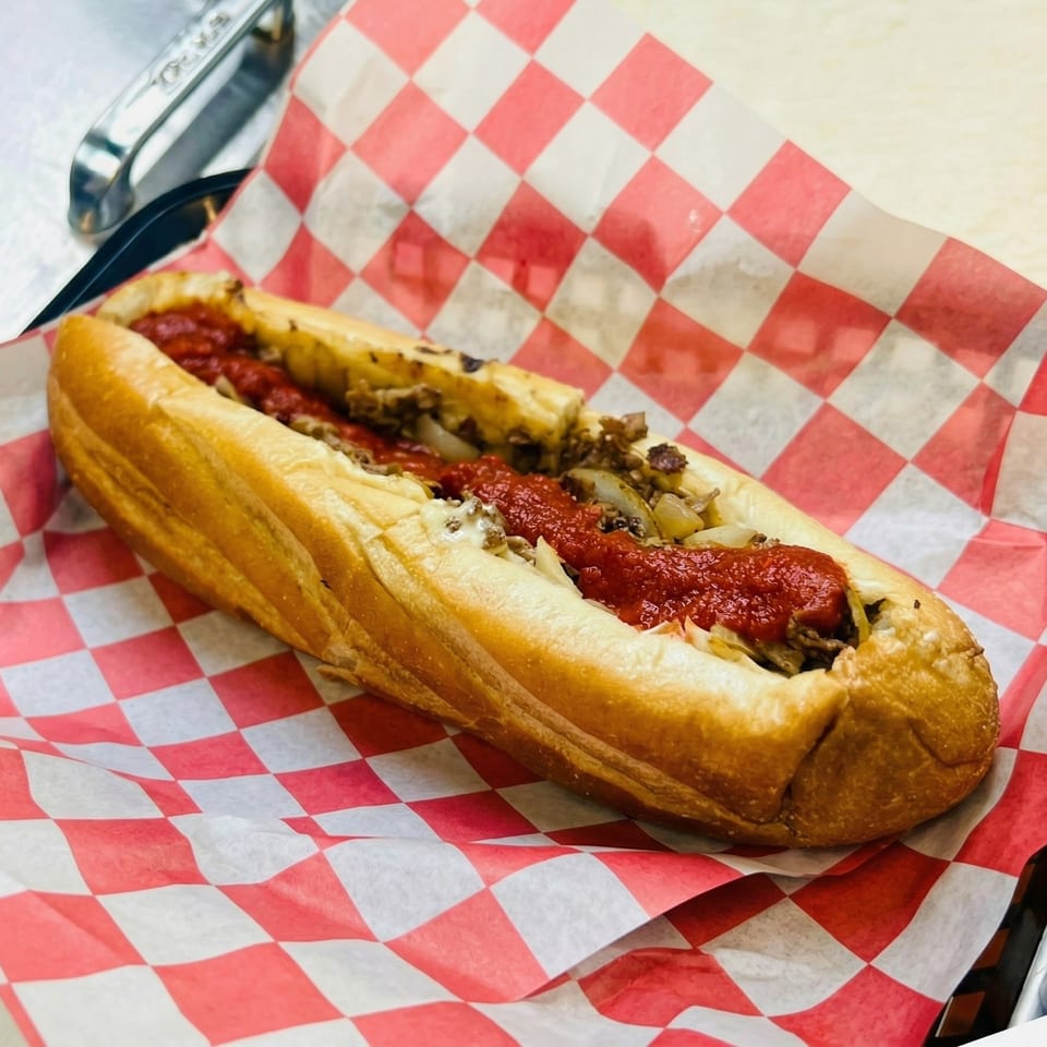 A delicious cheesesteak sandwich with red sauce and onions, served on red and white checkered paper.
