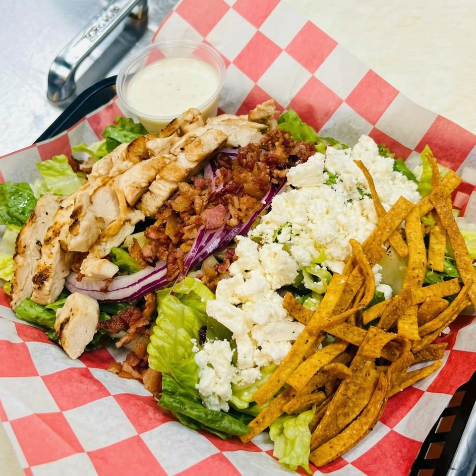 Grilled chicken salad with bacon, feta, red onion, and tortilla strips in a red checkered basket.